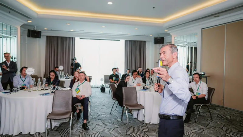 Giảng viên đứng thuyết trình trước các bàn tròn trong lớp CEO Bootcamp 2026, người tham dự đeo thẻ tên và tai nghe phiên dịch