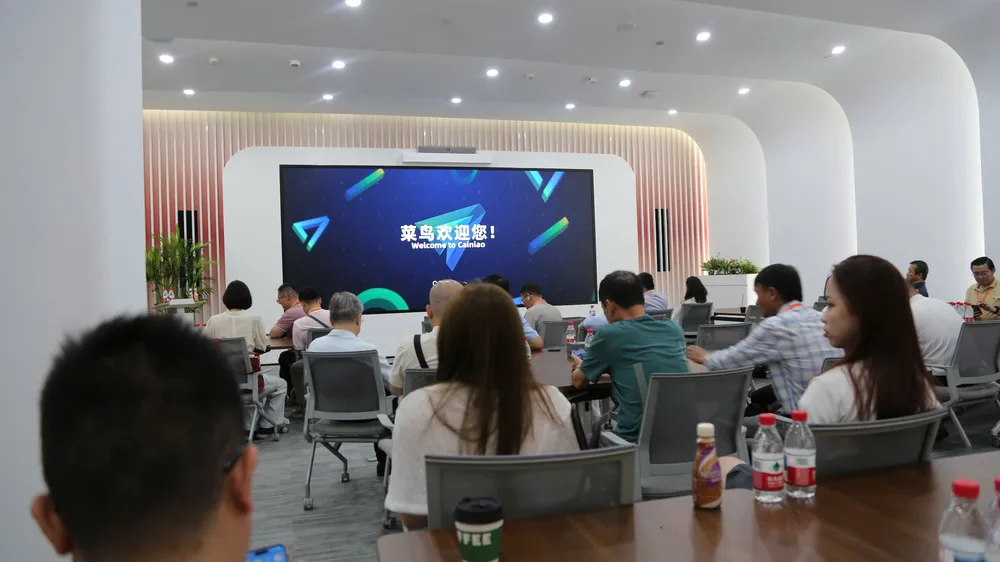 Đoàn Newing ngồi trong hội trường Cainiao, màn hình lớn hiển thị “菜鸟欢迎您！ Welcome to Cainiao”